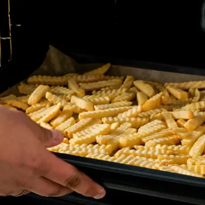 A hand sliding a dark baking sheet loaded with frozen crinkle-cut French fries into an oven