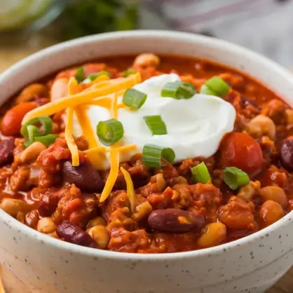A white bowl filled with thick chili, topped with a dollop of sour cream, shredded yellow cheese, and chopped green onions