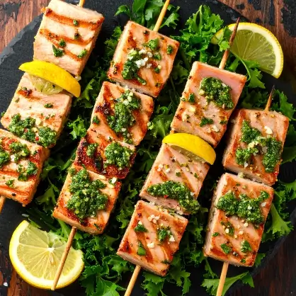 Grilled salmon pieces threaded onto wooden skewers, topped with a vibrant green herb sauce, and arranged on a bed of leafy greens with lemon slices