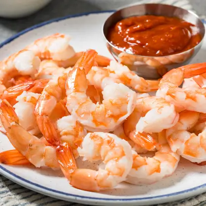 A white plate piled with cooked, chilled pink shrimp, accompanied by a small metal dipping bowl filled with red cocktail sauce