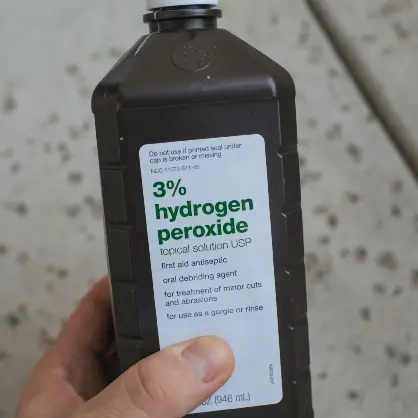 A hand holding a brown plastic bottle with a white label that reads "3% hydrogen peroxide topical solution USP"