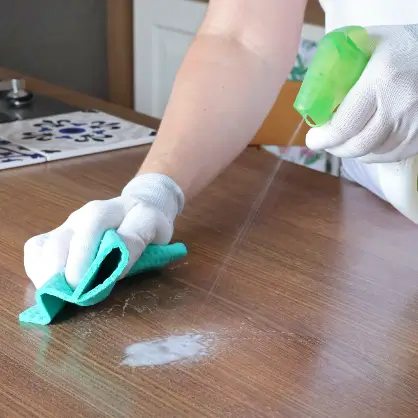 Gloved hands spraying cleaning liquid from a green bottle onto a wooden surface and wiping it down with a teal cloth