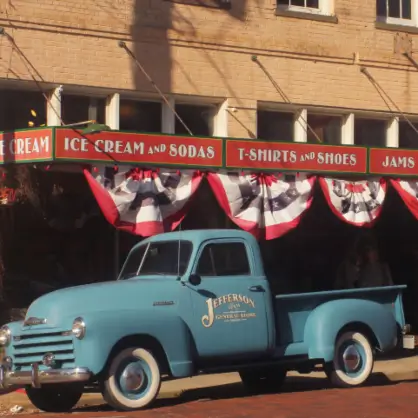 A vintage light blue pickup truck parked in front of a brick storefront with a red awning that reads "ICE CREAM AND SODAS" decorated with patriotic bunting.