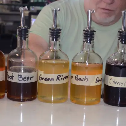 Several glass bottles with dispensing pumps containing different colored flavor syrups lined up at a counter, with labels like "Root Beer," "Green River," and "Peach."