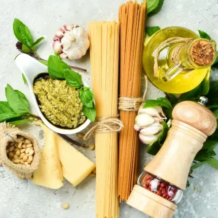 Pasta ingredients laid out on a table - spaghetti noodles, olive oil, seasoning, cheese, garlic, and vegetables