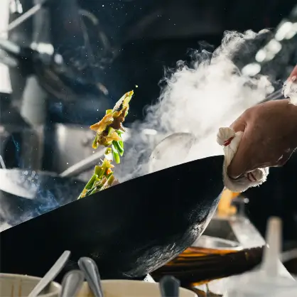 A chef actively tossing food in a wok over high heat, creating a large plume of steam and smoke