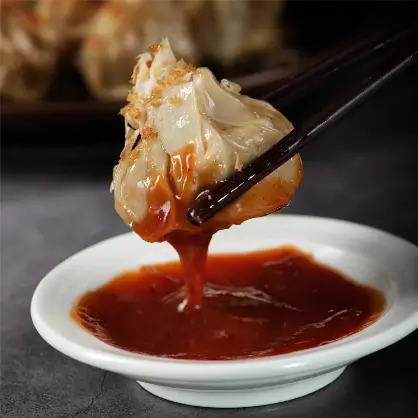Chopsticks holding a dumpling that is dripping a dark, reddish-brown sauce into a small white dipping bowl