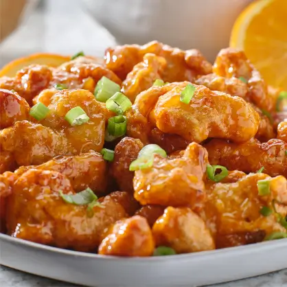 A bowl filled with pieces of fried, glossy orange chicken topped with chopped green onions, with an orange slice in the background