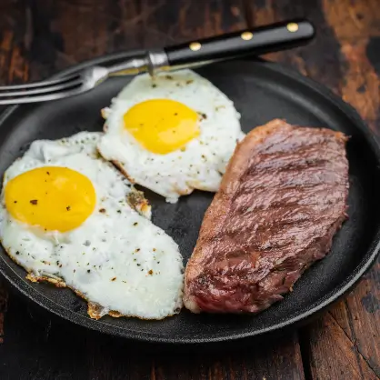 A black plate containing a cooked steak and two fried eggs sunny-side up, with a silver fork resting on the edge