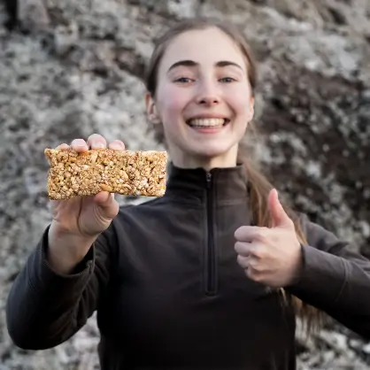 A smiling young woman outdoors holding up a rectangular granola bar in one hand while giving a thumbs-up with the other