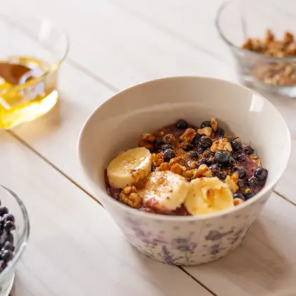 A white bowl filled with granola, fresh blueberries, and sliced bananas resting on a white wooden table