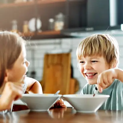 Two young children sitting together at a bright kitchen counter, smiling and happily eating breakfast from white bowls