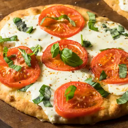 A close-up of a flatbread pizza topped with melted white cheese, sliced red tomatoes, and fresh green basil leaves
