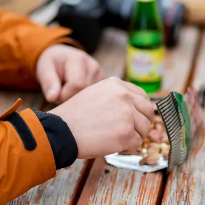 A close-up of a person's hands opening a small snack packet of trail mix on a rustic wooden table outdoors