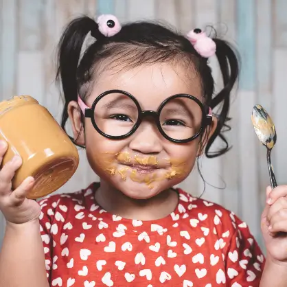 A smiling young girl with round glasses and peanut butter smeared on her face, happily holding a jar of peanut butter and a spoon