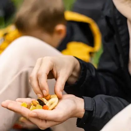 A close-up of an open hand holding a small handful of trail mix containing nuts and dried fruit, with a blurred person in the background