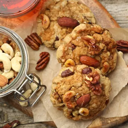 Several large, freshly baked cookies studded with whole nuts resting on parchment paper next to an open glass jar filled with mixed nuts