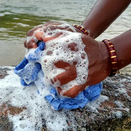 A close-up of a person's hands washing soapy blue fabric on a rocky surface near a body of water