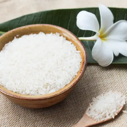 A wooden bowl overflowing with uncooked white rice, resting on a burlap cloth next to a white plumeria or jasmine flower on a green leaf