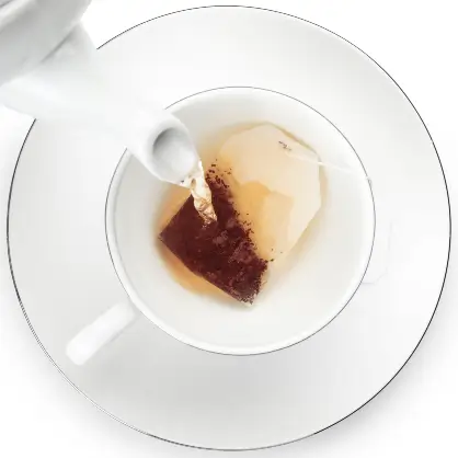 A top-down view of hot water being poured from a white teapot over a tea bag in a white cup