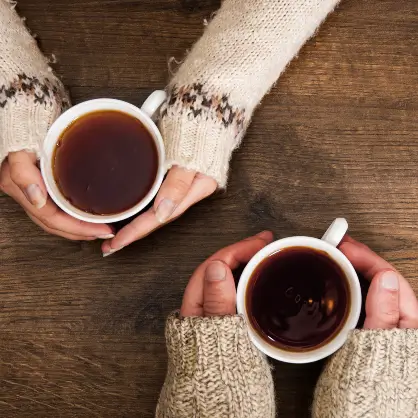 A top-down view of two people in cozy knit sweaters holding mugs of hot dark tea over a wooden table