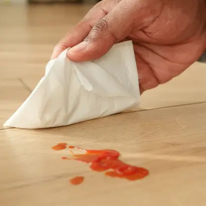 A hand holding a paper towel, about to clean up a red liquid spill on a wooden surface
