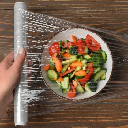 A hand pulling clear plastic wrap from a roll over a bowl of fresh vegetable salad