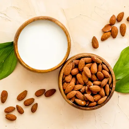 Top-down view of a white bowl filled with almond milk next to a wooden bowl full of whole almonds, with scattered almonds and green leaves on a beige background