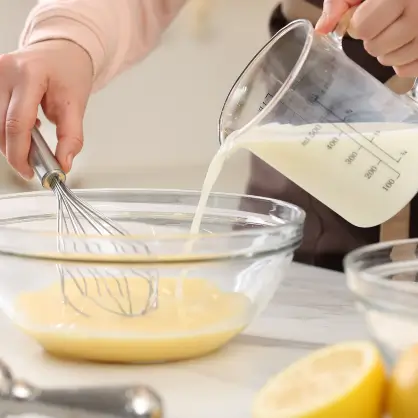 A person pouring a liquid from a glass measuring cup into a glass bowl while holding a whisk, with half a lemon resting on the counter nearby