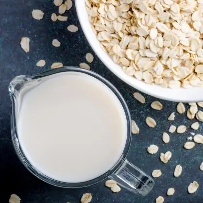 Top-down view of a glass pitcher filled with oat milk surrounded by scattered rolled oats and a bowl of oats on a dark surface