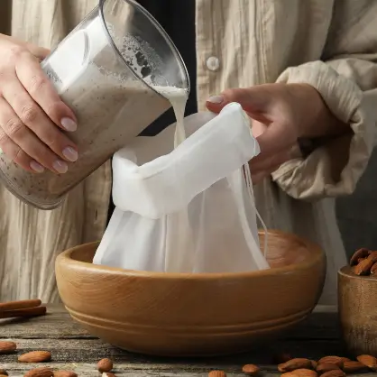 Hands holding a white straining bag over a wooden bowl while pouring a blended nut mixture from a glass pitcher to filter the milk