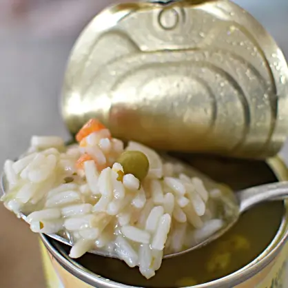 A close-up of a spoon lifting a scoop of thick rice and vegetable soup directly from an open tin can