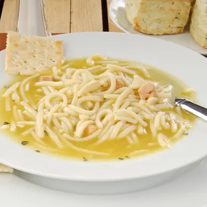 A shallow white bowl filled with chicken noodle soup, with a single square saltine cracker resting on the rim