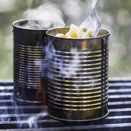 Two open, ridged metal cans heating on a grill grate with steam rising, as a fork lifts pasta out of the front can