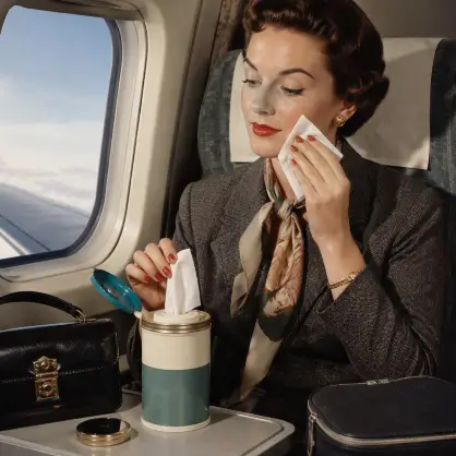 A woman in 1950s attire sitting by an airplane window, pulling a wet wipe from a cylindrical container on her tray table to clean her face