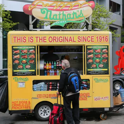 A bright yellow Nathan's Famous hot dog food cart parked on a city street with a customer wearing a backpack standing in front