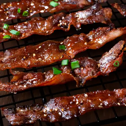 Strips of cooked, crispy bacon resting on a black wire cooling rack, lightly garnished with small green scallions