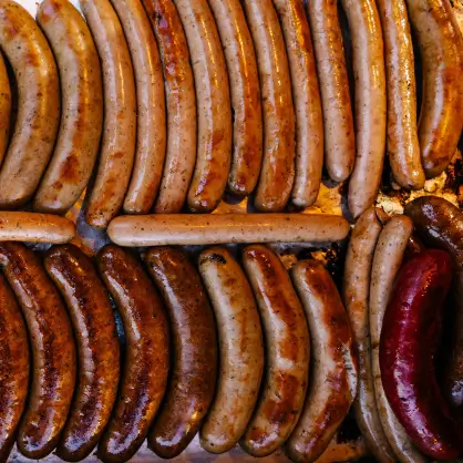Dozens of bratwursts and sausages of varying sizes and shades of brown cooking close together on a grill
