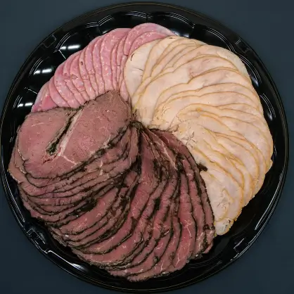 A round black platter neatly arranged with overlapping, circular fan patterns of sliced deli meats including roast beef, turkey, and ham