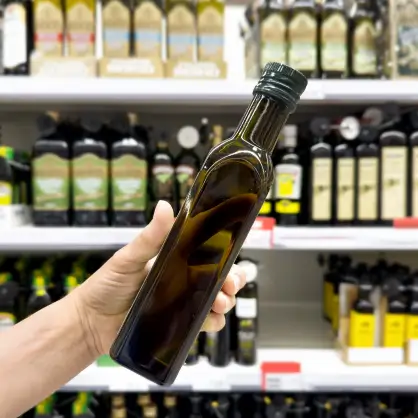 A hand holding up a dark square glass bottle of olive oil in front of blurred supermarket shelves stocked with various cooking oils
