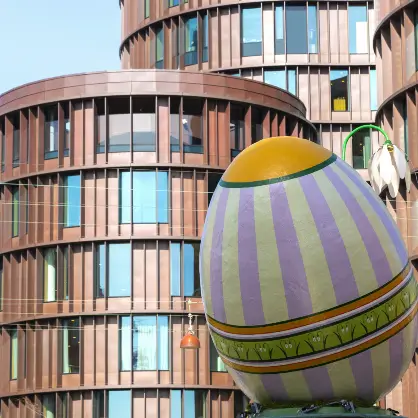 A massive, colorful decorated Easter egg sculpture with purple and yellow stripes standing outdoors in front of a modern, curved building