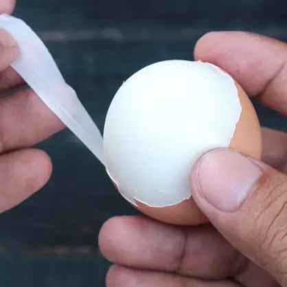 A close-up of a person's hands gently peeling the cracked brown shell off of a smooth, white hard-boiled egg