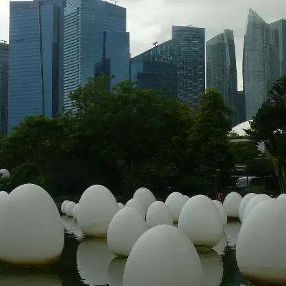 Numerous large, white, egg-shaped sculptures floating on a body of water in an urban park, with modern city skyscrapers towering in the background